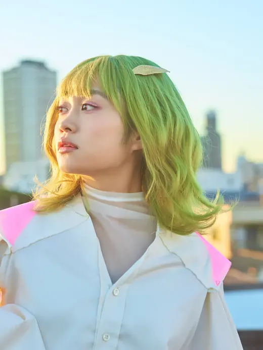 緑の髪に葉のヘアピンをつけ、夕暮れの街を背に横を向くアートの子